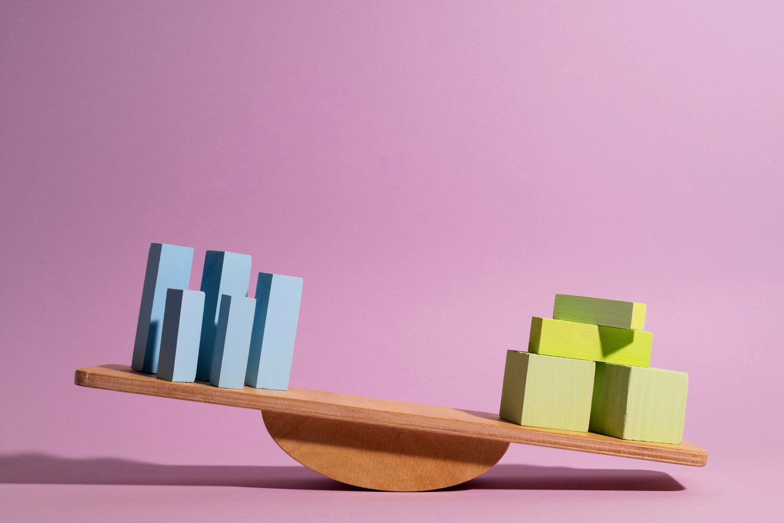 Een houten wip staat op een roze achtergrond uit balans. Links staan meerdere slanke, rechtopstaande blokken in tinten lichtblauw en grijsblauw — ze wijzen omhoog, maar het uiteinde van de wip is omhooggekanteld. Rechts ligt het zwaarste gewicht: een piramide van brede, gestapelde blokken in geelgroen, die de wip naar beneden trekt. Het beeld visualiseert een verstoord evenwicht tussen twee kanten die niet gelijkwaardig wegen.